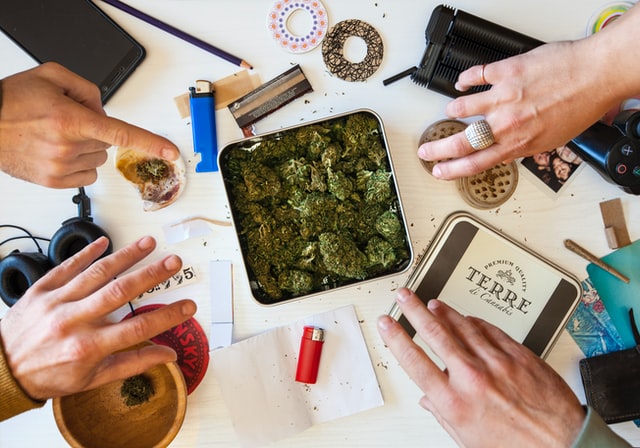 Hands surrounding a table covered with cannabis buds in a metal tin, grinder, lighter, rolling papers, and various accessories.