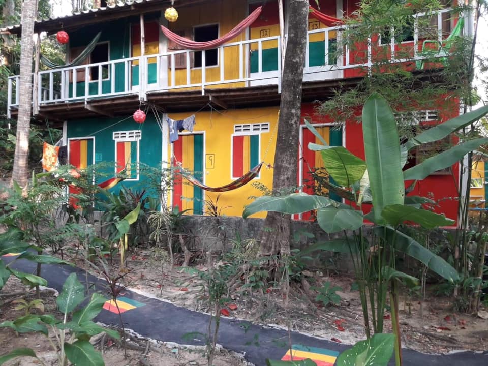 Two-story colorful building with green, yellow, and red painted walls and hammocks hanging on the balcony, surrounded by lush vegetation.