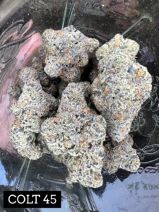 Several cannabis buds in a glass jar. The buds are gray, green, and purple, and the words 