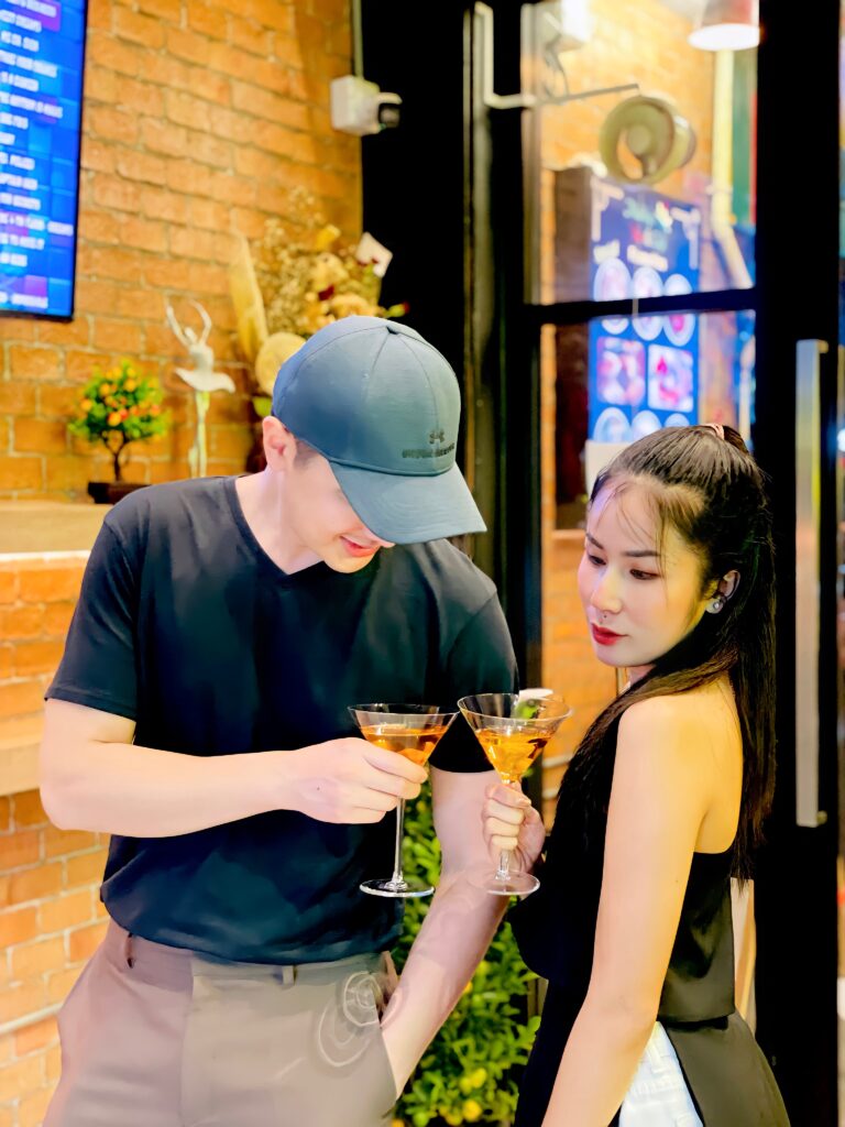 A man and woman hold cocktail glasses in a dimly lit bar setting, toasting each other.