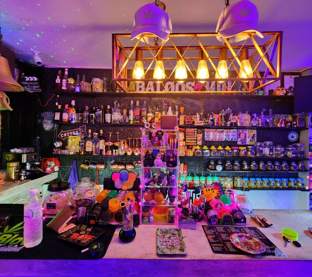 Bar interior with shelves displaying bottles and jars. Two caps with a cannabis leaf are visible. The word 