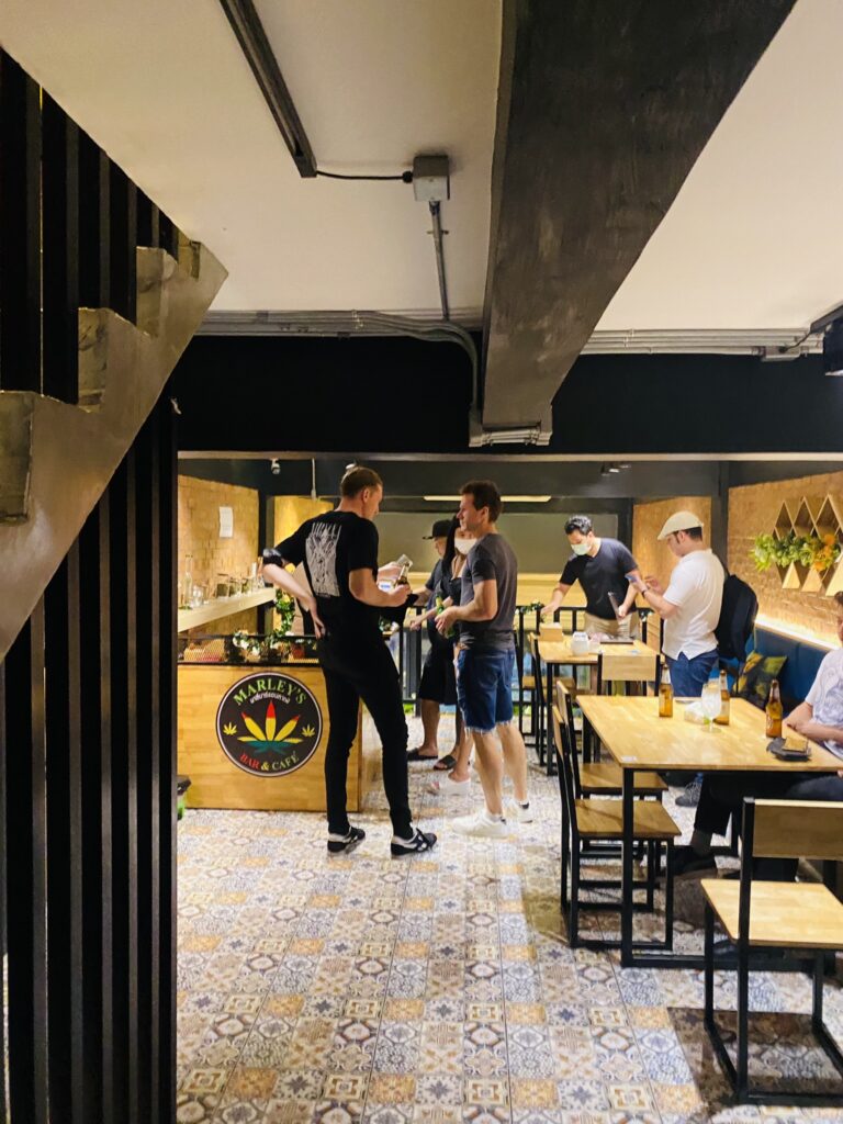 People at tables and standing around in Marley's Bar & Cafe, wooden tables, patterned tile floor, and a bar with a cannabis leaf logo.