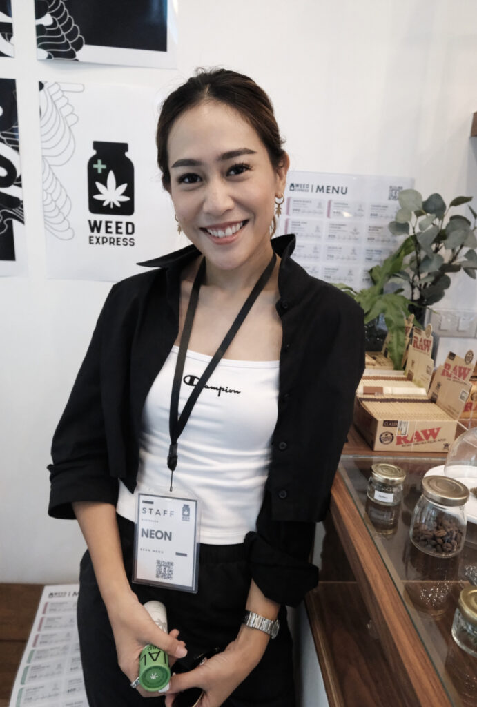 Woman smiling, wearing a black jacket, white top, and STAFF badge, standing in front of a WEED EXPRESS banner.