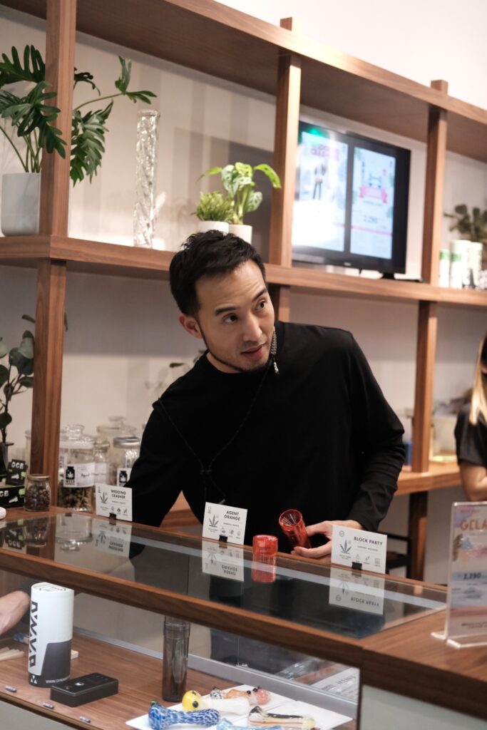 Man in black shirt standing behind a display counter, holding a red container. Shelves behind him hold plants and jars with labels like 