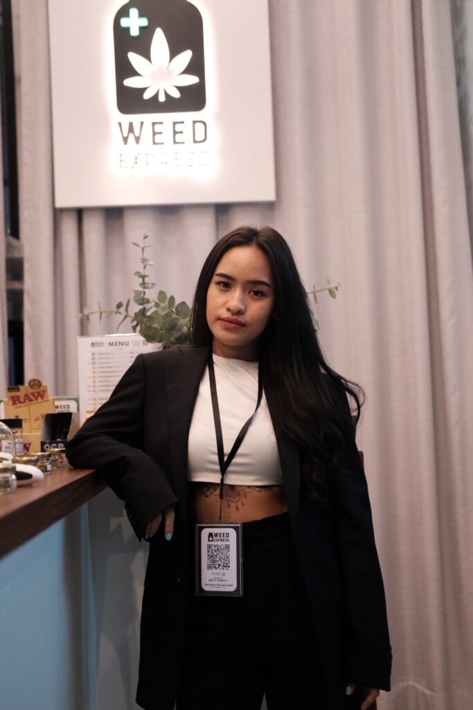 Woman with long dark hair wearing a black blazer, white crop top, and black pants, standing behind a counter in front of a 