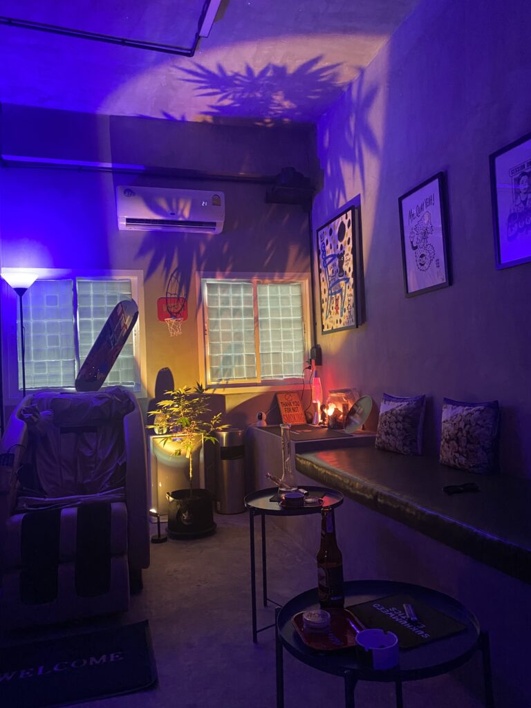 A dimly lit interior with purple lighting, a massage chair, a potted plant, a bong, a skateboard, and framed art on the wall.