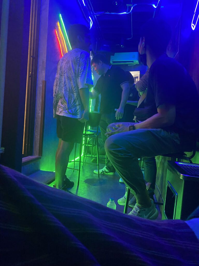 People in a bar with neon lighting; one person sits on a stool, and two others stand.