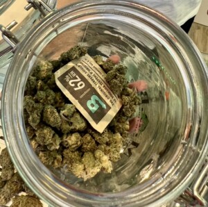 Glass jar filled with cannabis buds, a product label with 62% RH, and the letter 