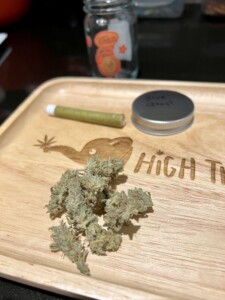Cannabis buds on a wooden tray with a pre-rolled joint, a lid labeled 