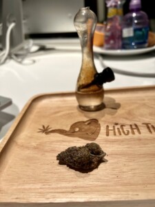 A glass water pipe rests on a wooden tray with a nugget of cannabis and the words 
