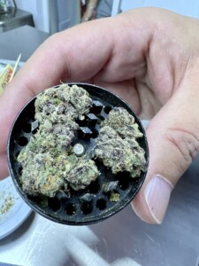 Hand holding a black grinder containing cannabis buds, with a plate and rolling papers visible in the background.