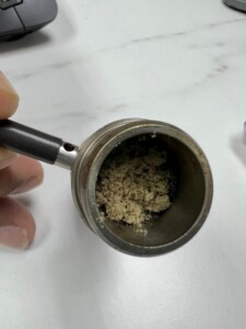 An open, metal part of a device filled with a tan, grainy substance is held by a hand on a white surface.