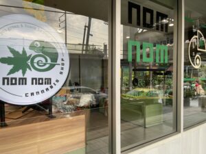 Nom Nom Cannabis Shop window, sign with a cannabis leaf and logo, reflection of street and car.