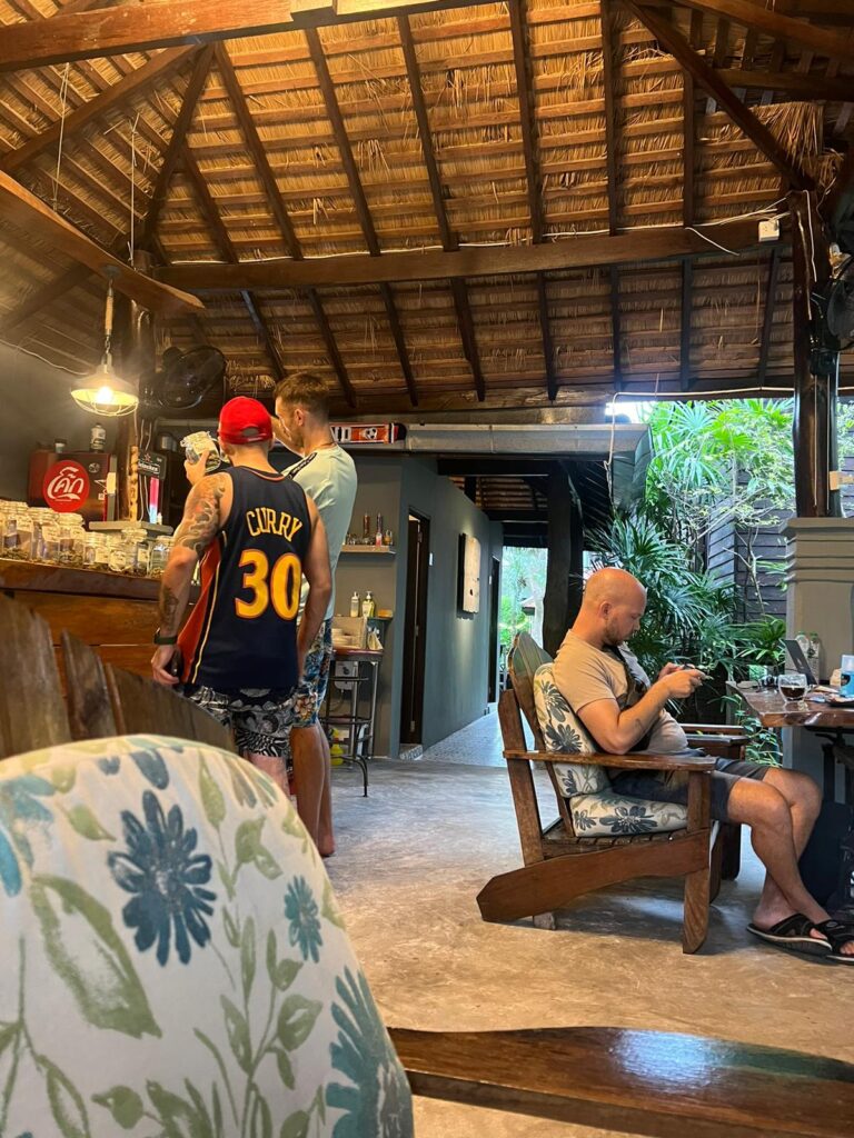 Three men inside a bar, one wearing a Curry 30 jersey, others in casual clothing. One man is sitting and looking at his phone. Bar has thatched roof.