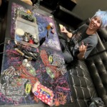 A person with blue hair on a black leather sofa smiles, making a peace sign. A table with marijuana paraphernalia is visible.