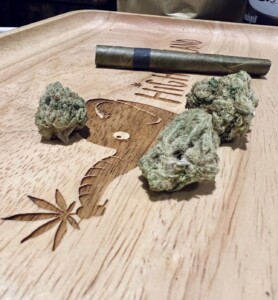 Rolled cannabis cigar and three buds on a wooden tray with etched marijuana leaf and elephant design.