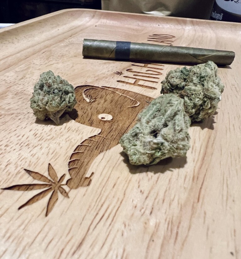 Rolled cannabis cigar and three buds on a wooden tray with etched marijuana leaf and elephant design.