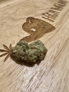 A light green cannabis bud on a wooden surface with a carved cartoon character and the words 