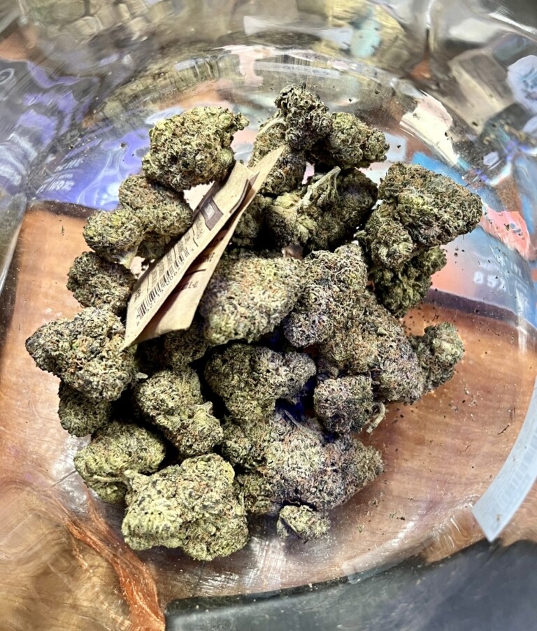 Green and purple cannabis buds filling a clear glass jar, with a folded brown paper tag visible.