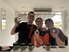 Happy customers at Blessed Ayutthaya, Thailand, showing cannabis flower jars. Strains: Dirty Bird, Apple Fritter, and more.