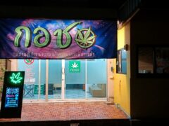 Exterior of Kor Chor cannabis shop, showing a banner and neon open sign, many strains of weed available.