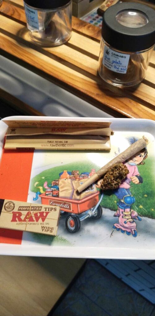 Rolling tray with a lit cannabis joint, cannabis flower, Raw rolling papers, and a jar labeled 