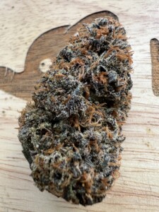 A cannabis bud with dense, purple and orange trichomes rests on a light wood surface.
