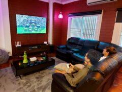 Two people relaxing on a black leather couch watching TV, with a green decorative object on the black coffee table.