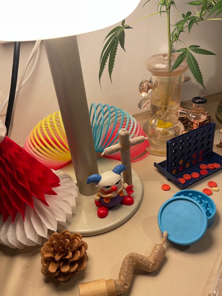 Still life: lamp, toys, cannabis leaves in vase, connect four, pinecone, and joint.
