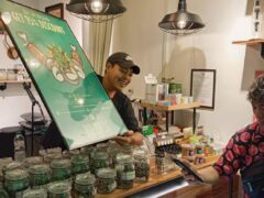 HARMONY HERB dispensary in Pattaya, with cannabis flower jars, a tattoo shop, and customer scanning QR code for a discount.