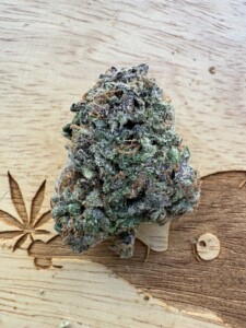 Bud of green and purple cannabis with trichomes on a wooden surface, a carved marijuana leaf is on the left.