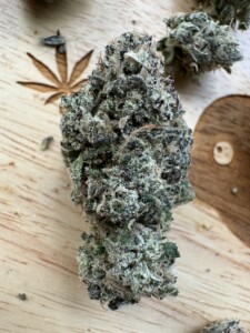 A dense, green and frosty cannabis bud on a wooden surface, with a cannabis leaf design and another bud visible.