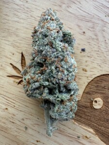 Cannabis bud with green, purple, and orange coloring on a wooden surface with carved leaf and circle.
