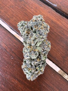 A large, dense cannabis bud with green, purple, and orange highlights, resting on a dark brown wooden surface.