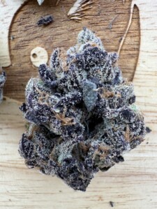 A close-up of a cannabis bud, heavily covered with trichomes, set on a wood surface. The bud has dark purple and green hues.
