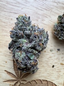 A close-up of cannabis bud with purple and green hues, with small crystals, on a wooden surface with a carved leaf design.