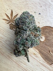 A dense cannabis bud, with green, purple, and brown hues, rests on a wooden surface with a carved cannabis leaf detail.