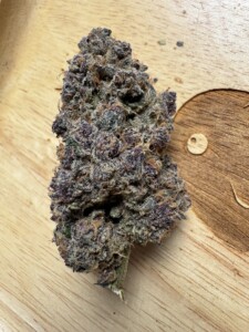 A close-up of a cannabis bud on a wooden surface. The bud is dense and covered in trichomes, with visible purple and green hues. The wooden surface has a natural wood grain.