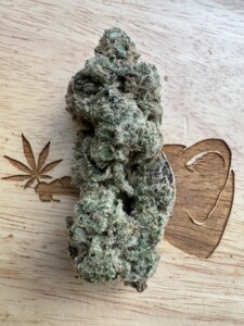 Green cannabis bud with white trichomes, on a wooden surface with carved hemp leaf and heart shapes.