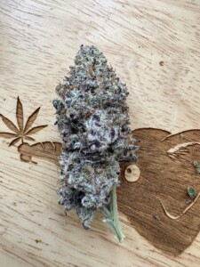 A dense, purple and green cannabis bud on a wooden surface with carved cannabis leaf.