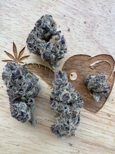Four cannabis buds on a wooden surface, with a leaf and heart-shaped cutout.