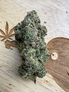 Green and frosty cannabis bud on a wooden surface, next to a carved cannabis leaf.