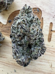 A close-up of a marijuana bud with frosty white trichomes and purple and green hues, resting on a wooden surface.
