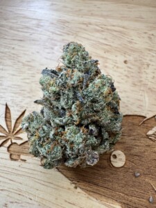 A close-up of a cannabis bud, showing green, purple, and orange plant material on a wooden surface.