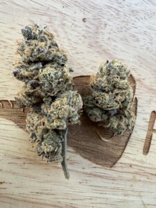 Two cannabis buds on a wooden surface with a partial carving of a leaf.
