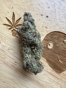 A cannabis bud on a wooden surface with a carved leaf and a smiling face.