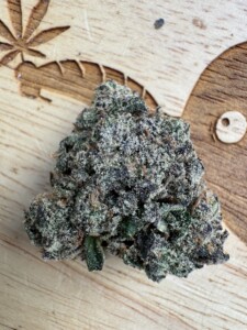 A close-up of a cannabis bud with purple, green, and white trichomes on a wooden surface.
