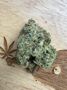 A green cannabis bud rests on a wooden surface with a carved marijuana leaf.