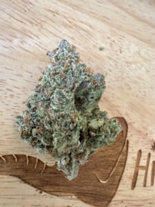 A green and frosty cannabis bud on a light wood surface with an etched design.
