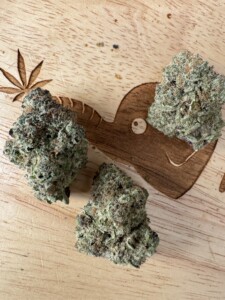 Three cannabis buds on a light wood surface, a carved leaf design, and a carved heart with a dot inside.